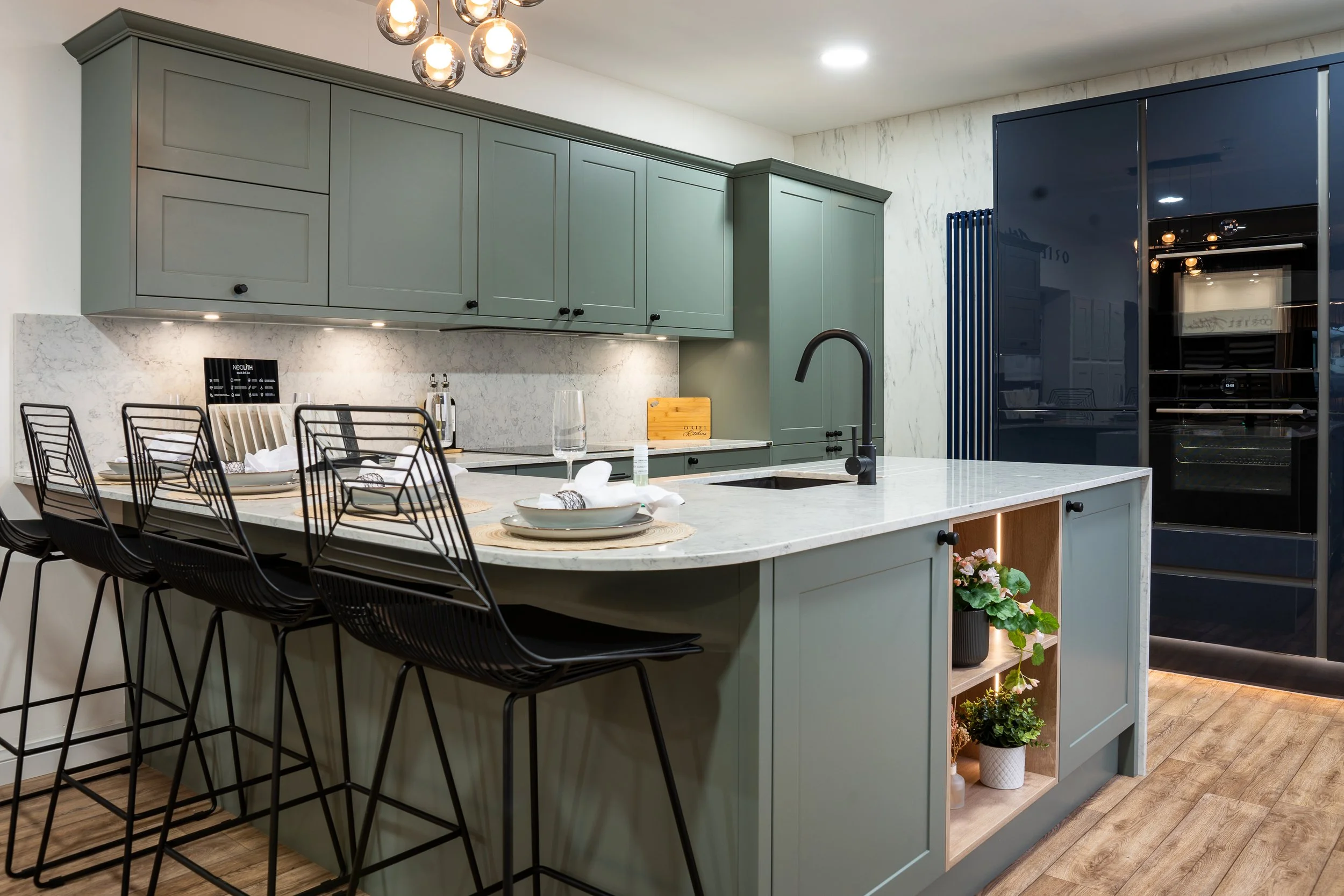 The sage-green shaker peninsula in the Oriel Kitchens showroom at Lion House, Abergavenny, with a marble-quartz worktop, Belfast undermount sink and two navy-gloss tall ovens housings on the right