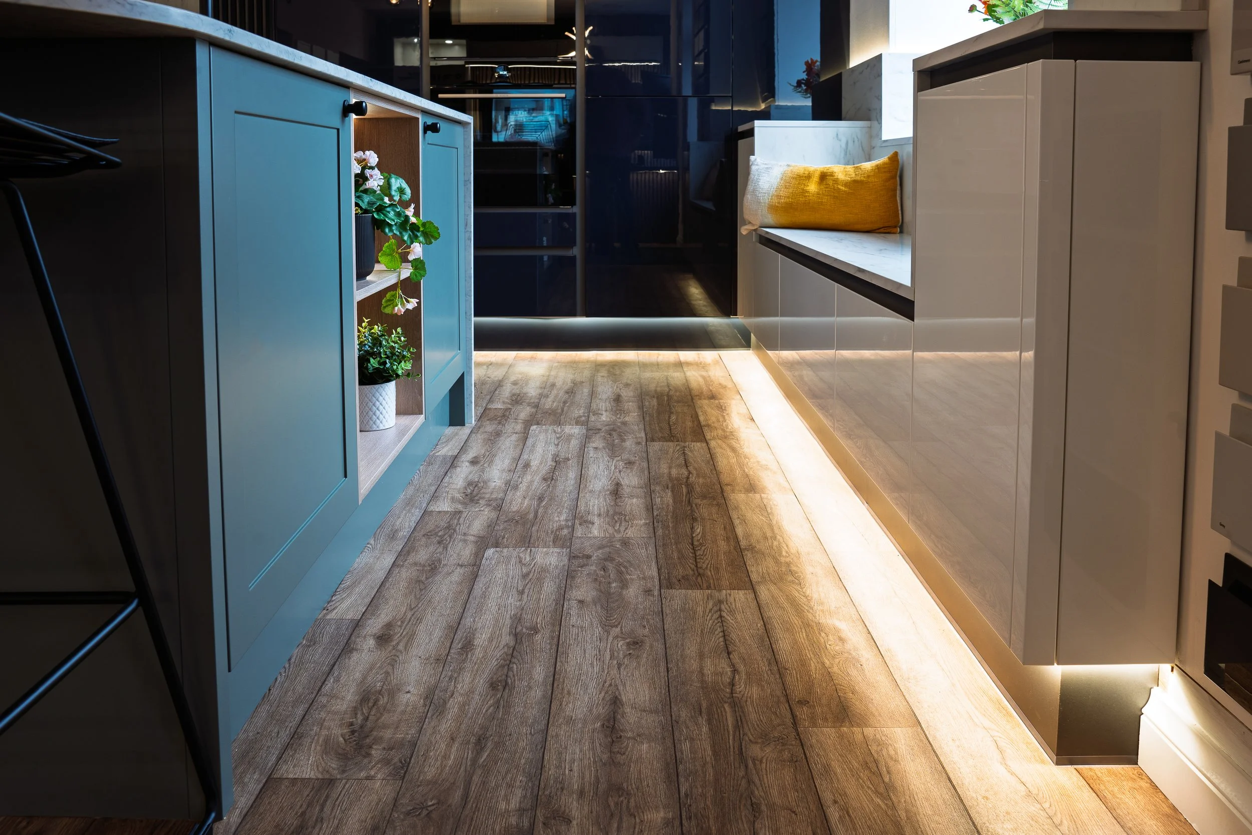 Low-angle showroom floor shot at Lion House: sage-green shaker peninsula on the left with an open display cabinet and a trailing pothos, deep navy gloss panel on the right reflecting LED-strip floor lighting, real oak-look LVT plank floor