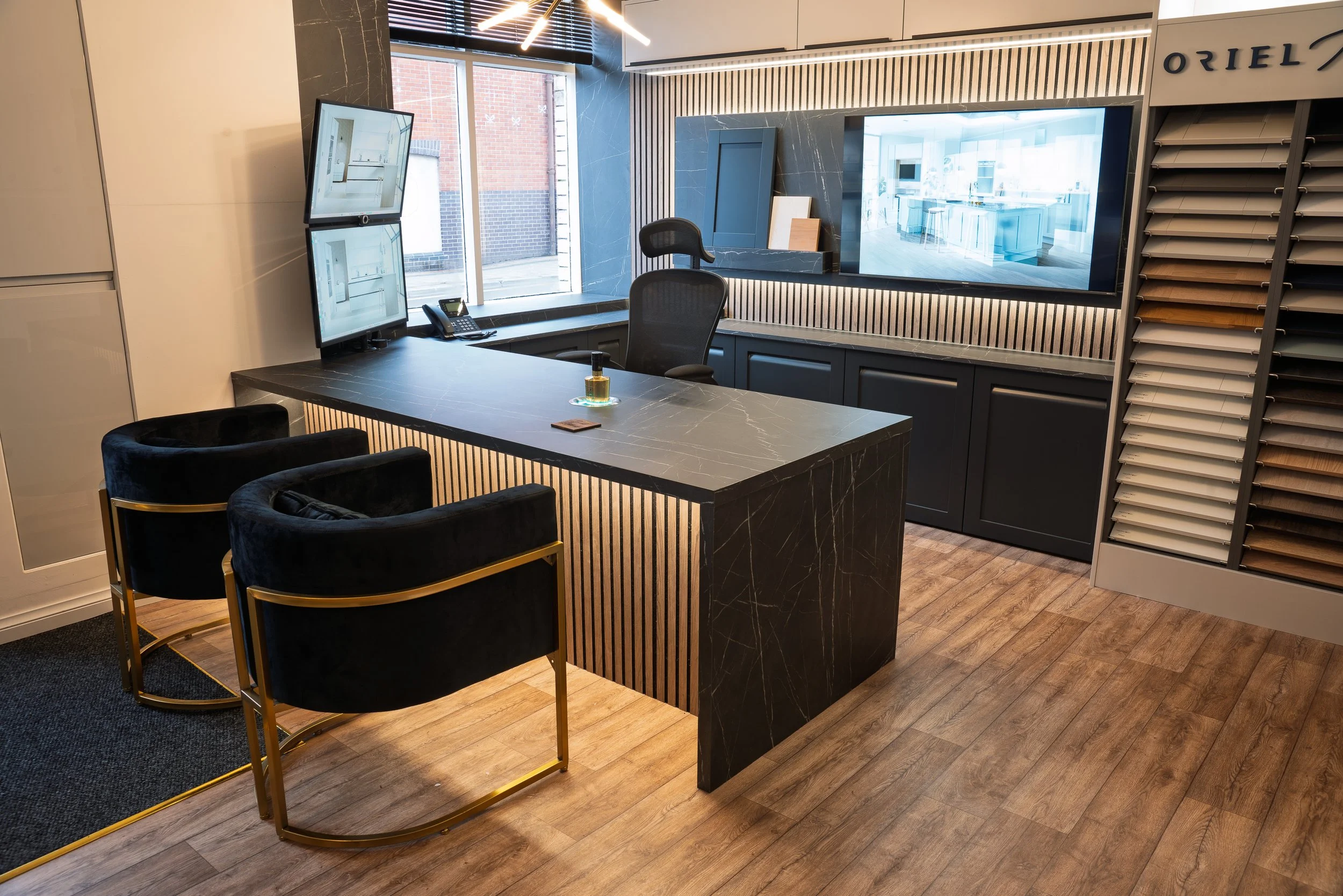The design consultation desk inside the Lion House showroom: black slatted timber wall behind, dark Calacatta-marble desk, two black Eames-style swivel chairs, dual monitors showing CAD plans, the ORIEL wordmark on the wall-mounted door-sample rack to the right